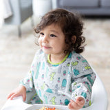 A toddler in a Bumkins Sleeved Bib: Cacti sits in a high chair with a fork in hand, ready to eat.