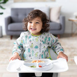 A toddler in a Bumkins Sleeved Bib: Cacti enjoys spaghetti from a plate in a high chair with a sofa and side table visible in the background.