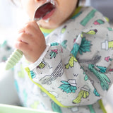 Close-up of a baby holding a food-covered spoon, wearing a Bumkins Sleeved Bib: Cacti.