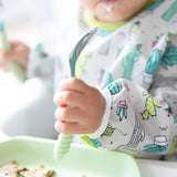 Close-up of a baby wearing a Bumkins Sleeved Bib: Cacti, holding a green spoon over a green plate.