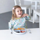 A smiling child wearing a Bumkins Sleeved Bib: Cacti sits at a white table with fruit and pasta on a gray plate near gray utensils.