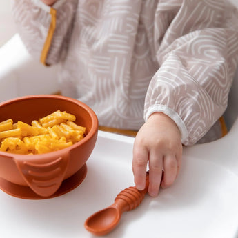 Close-up of a child in a Bumkins Sleeved Bib: Wander reaching for a spoon beside a bowl of mac and cheese on a high chair tray.