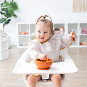 A toddler wearing a Bumkins Sleeved Bib: Wander smiles while eating from an orange bowl at a high chair; toys fill shelves behind.