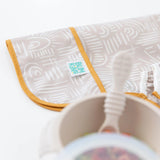 Close-up of a folded Bumkins Sleeved Bib: Wander on a white background, with a blurred bowl and spoon in the foreground.