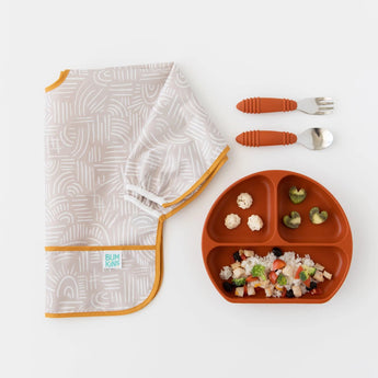 Bumkins Sleeved Bib: Wander is folded on a white background, next to a dark orange divided plate of food and utensils.