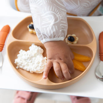 Close-up of a child's hand reaching for orange slices on a plate with cottage cheese and snacks, while wearing a Bumkins Sleeved Bib: Wander.