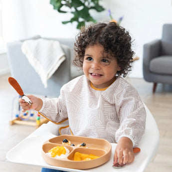 A curly-haired toddler grins in a high chair, clutching a spoon and wearing a Bumkins Sleeved Bib: Wander as they eat from a tan dish.