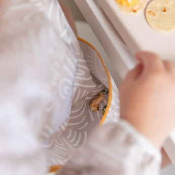 Close-up of a child in a Bumkins Sleeved Bib: Wander with food in the pocket, sitting at a table with tortilla pieces.