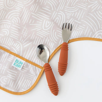 Close-up of a spoon and fork with orange rubber handles on top of a Bumkins Sleeved Bib: Wander on a white background.