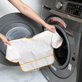 A person holding a Bumkins Sleeved Bib: Wander in front of an open front-loading washing machine.