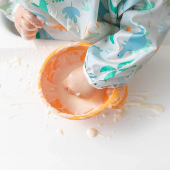 Close-up of a child's hand playing with yogurt in an orange bowl, wearing a Bumkins Sleeved Bib: Dinosaurs.