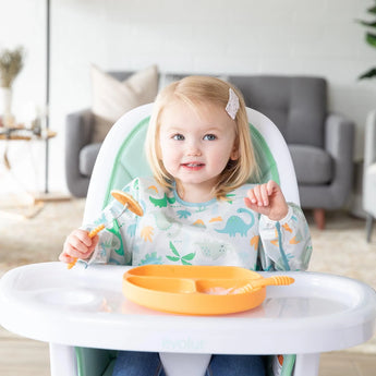 A toddler in a high chair, wearing a Bumkins Sleeved Bib: Dinosaurs holds a fork with a pancake near an orange plate.