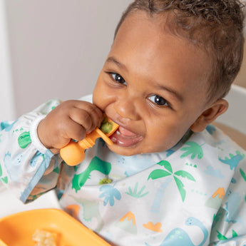 A smiling baby joyfully eats with an orange spoon and plate, wearing a Bumkins Sleeved Bib: Dinosaurs.