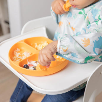 Close-up of a toddler in a high chair wearing a Bumkins Sleeved Bib: Dinosaurs, eating from an orange divided plate with a spoon.
