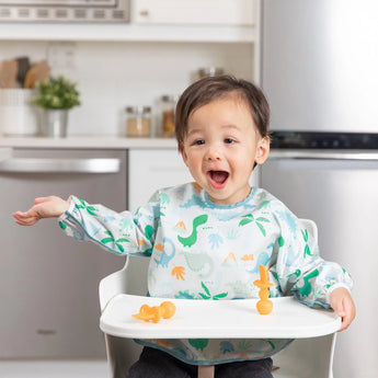 A joyful toddler in a high chair, wearing a Bumkins Sleeved Bib: Dinosaurs, raises their arm excitedly, with orange utensils on the tray.