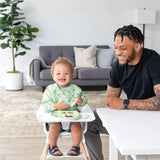 A joyful toddler enjoys mealtime in a Bumkins Sleeved Bib: Llamas, as a tattooed man sitting at a white table smiles and watches.