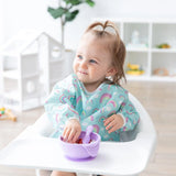 A toddler in a high chair grins while eating from a purple bowl, wearing a Bumkins Sleeved Bib: Rainbows in a bright room.