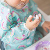 Close-up of a toddler covered in yogurt, wearing a Bumkins Sleeved Bib: Rainbows.