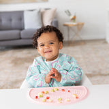 A curly-haired toddler eats fruit from a pink silicone mat, wearing a Bumkins Sleeved Bib: Rainbows in a bright living room.