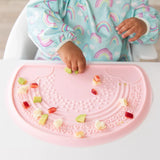 Close-up of a baby in a Bumkins Sleeved Bib: Rainbows playing with food on a pink silicone placemat.