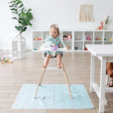 A toddler in a high chair wears a Bumkins Sleeved Bib: Rainbows, eating from a purple bowl in a bright playroom with toys.