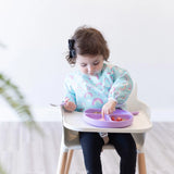 A toddler wears a Bumkins Sleeved Bib: Rainbows, eating fruit with a spoon from a purple plate in a high chair.