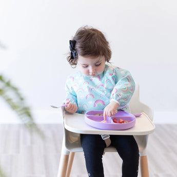 A toddler wears a Bumkins Sleeved Bib: Rainbows, eating fruit with a spoon from a purple plate in a high chair.
