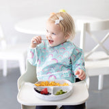 A toddler in a high chair, wearing a Bumkins Sleeved Bib: Rainbows, happily enjoys pasta, strawberries, and broccoli from a gray plate.