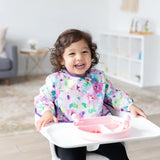 A toddler wearing a colorful bib smiles in a high chair with Bumkins Grip Dish Essential Infant 4-Piece Mealtime Set: Pink on the tray.