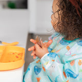 A curly-haired child in a Bumkins Sleeved Bib: Ocean Life holds a messy fork next to an orange plate with food remnants.