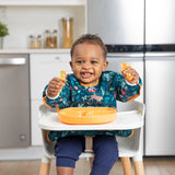A smiling toddler in a high chair, wearing Bumkins Sleeved Bib: Jungle, holds orange utensils near an orange divided plate.