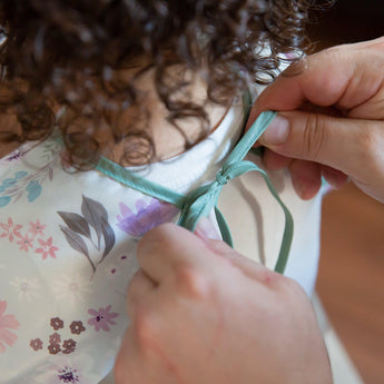Hands tying the ribbon closure of a Bumkins Sleeved Bib: Floral around a child's neck.