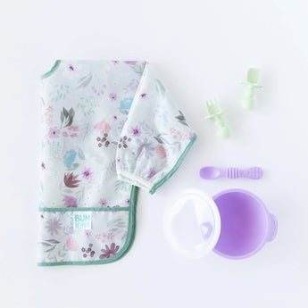 Bumkins Sleeved Bib: Floral lays folded on a white background next to a purple bowl and spoon set with lid, and green toddler utensils.
