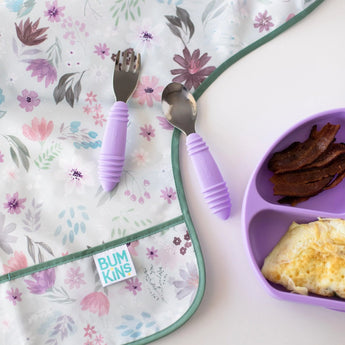 Close-up of Bumkins Sleeved Bib: Floral on a white background, with purple utensils on top and a purple plate of bacon and eggs nearby.