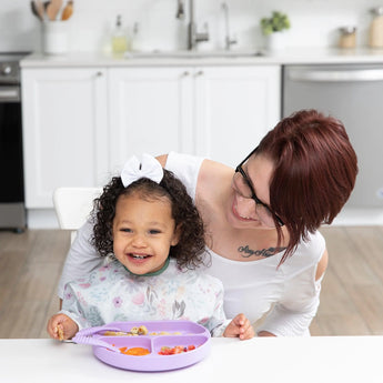 A smiling woman and toddler enjoy a meal at a white table; the toddler wears a white bow and a Bumkins Sleeved Bib: Floral.
