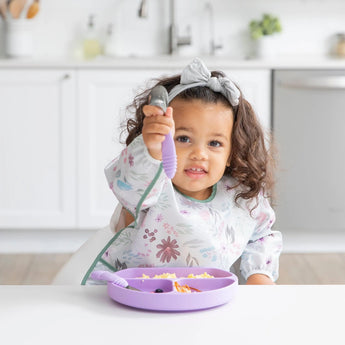 A toddler excitedly holds a spoon while wearing a Bumkins Sleeved Bib: Floral, sitting at a white table with a divided purple plate.