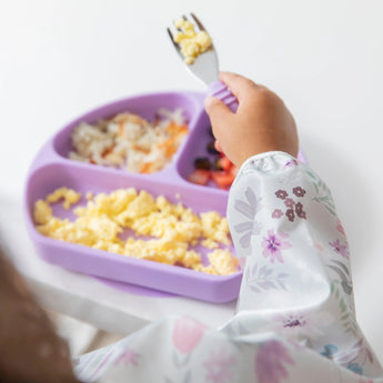 Close-up of a child's arm in Bumkins Sleeved Bib: Floral, holding a fork with scrambled eggs while eating from a purple plate.