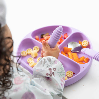 Close-up of a child in a Bumkins Sleeved Bib: Floral enjoying colorful foods from a purple divided plate with utensils at a white table.