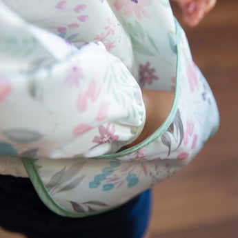 Close-up of a child wearing a Bumkins Sleeved Bib: Floral, made from waterproof fabric, with their hand tucked in the front pocket.