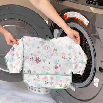 A person holds Bumkins Sleeved Bib: Floral in front of an open front-loading washing machine.
