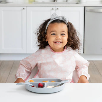 A young girl with curly hair and gray bow wears a Bumkins Sleeved Bib: Lace, sitting at a white table with fruit on a gray divided plate.