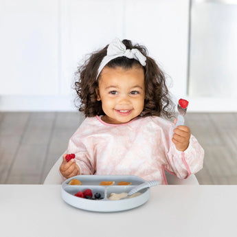 A curly-haired toddler in a Bumkins Sleeved Bib: Lace smiles, holding a fork with fruit, seated at a table with a divided plate of food.