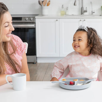 A woman and girl wearing a Bumkins Sleeved Bib: Lace smile at each other at a white table in a bright kitchen; a divided plate and cup nearby.