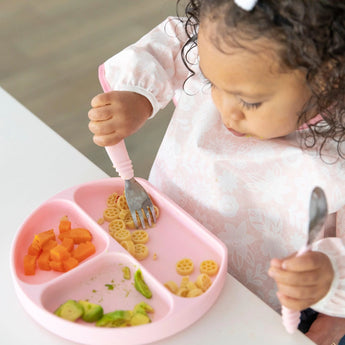A toddler enjoys pasta, carrots, and avocado from a pink plate while using utensils and wearing a Bumkins Sleeved Bib: Lace.