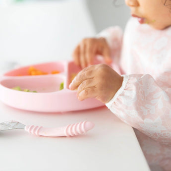 Close-up of a toddler in a Bumkins Sleeved Bib: Lace eating from a pink divided plate at a white table.