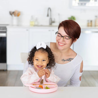 A woman in glasses watches a child in a Bumkins Sleeved Bib: Lace eat from a pink plate using a fork at a white table.