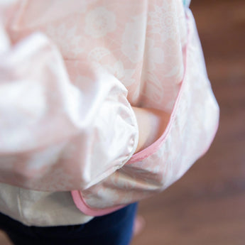 Close-up of a child wearing a Bumkins Sleeved Bib: Lace, with their hand in the pocket.
