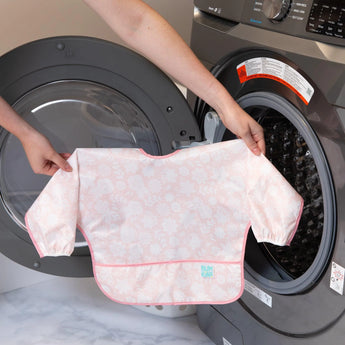 A person holds Bumkins Sleeved Bib: Lace in front of an open front-loading washing machine.