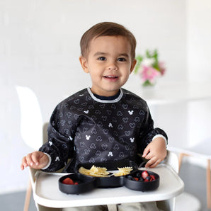 A toddler in a high chair, smiling with a divided plate of pasta and berries, wears a Bumkins Sleeved Bib: Mickey Mouse Icon Black + White.