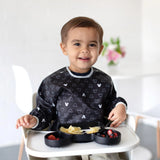 A toddler in a high chair, smiling with a divided plate of pasta and berries, wears a Bumkins Sleeved Bib: Mickey Mouse Icon Black + White.